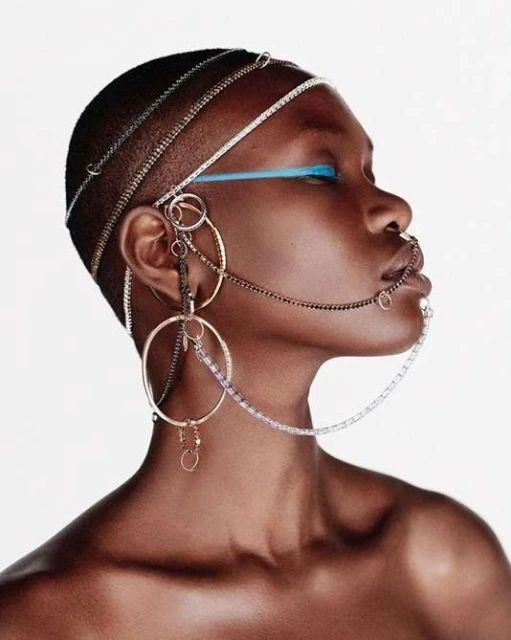 Profile portrait of a fashionable Black woman with short hair wearing distinctive gold and silver jewelry against a bright white backdrop.