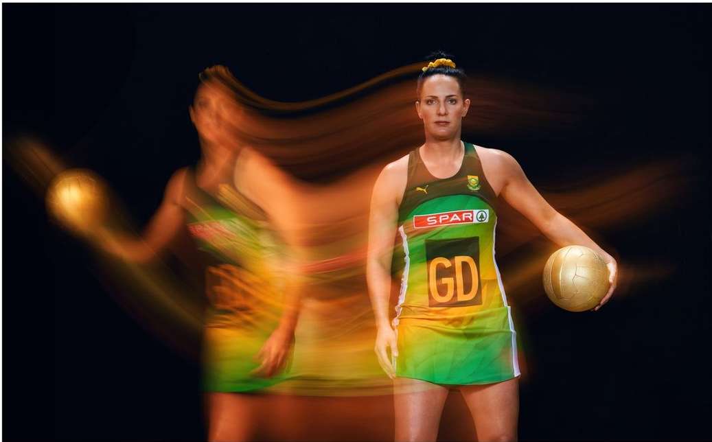 A woman in a netball uniform holds a ball. Another motion-blurred woman with a ball appears in the background against a black background.