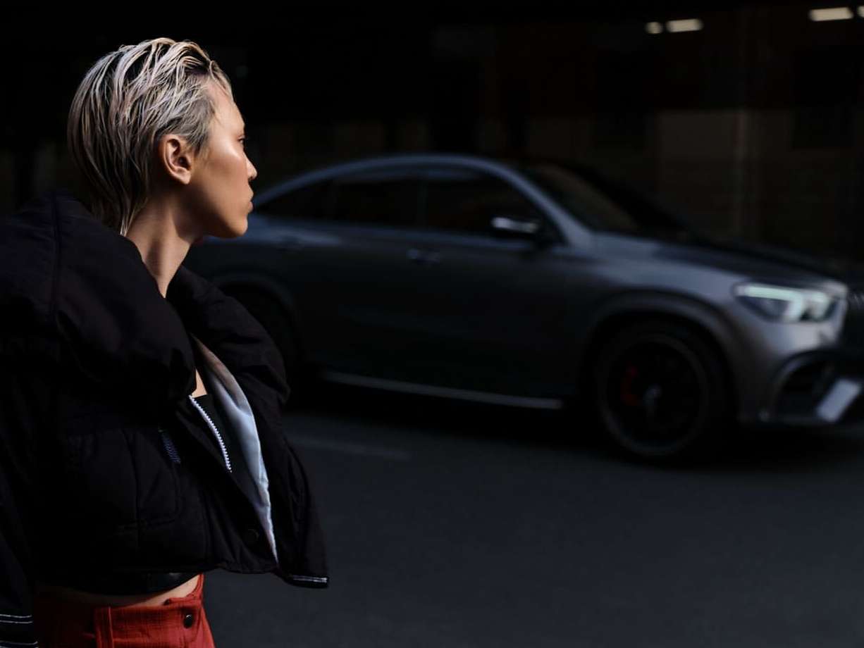Woman in a black jacket and red pants poses next to a dark grey SUV in an urban setting at dusk or dawn, with a shallow depth of field.