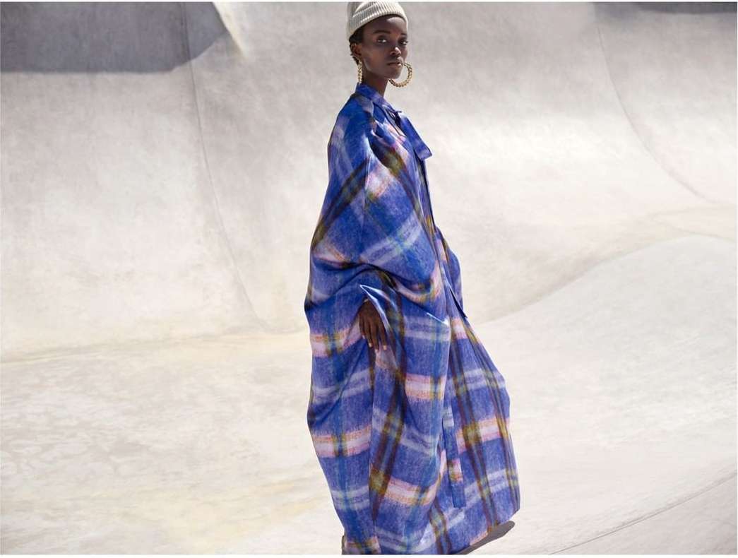 Fashion shot of a Black adult female model wearing a plaid blue coat and a beanie, posing in a concrete outdoor location with bright lighting.