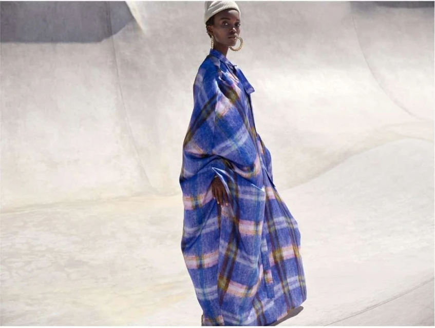 Fashion shot of a Black adult female model wearing a plaid blue coat and a beanie, posing in a concrete outdoor location with bright lighting.