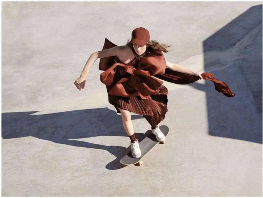 A young adult female in a brown dress and baseball cap skateboarding in a skatepark. She has white sneakers and neutral facial expression.