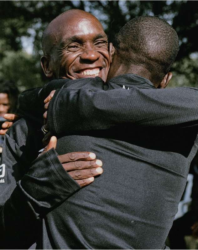 Two African-American men embracing outdoors, wearing black long-sleeved shirts, with one smiling broadly and the other partially obscured.
