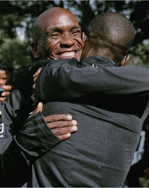 Two African-American men embracing outdoors, wearing black long-sleeved shirts, with one smiling broadly and the other partially obscured.