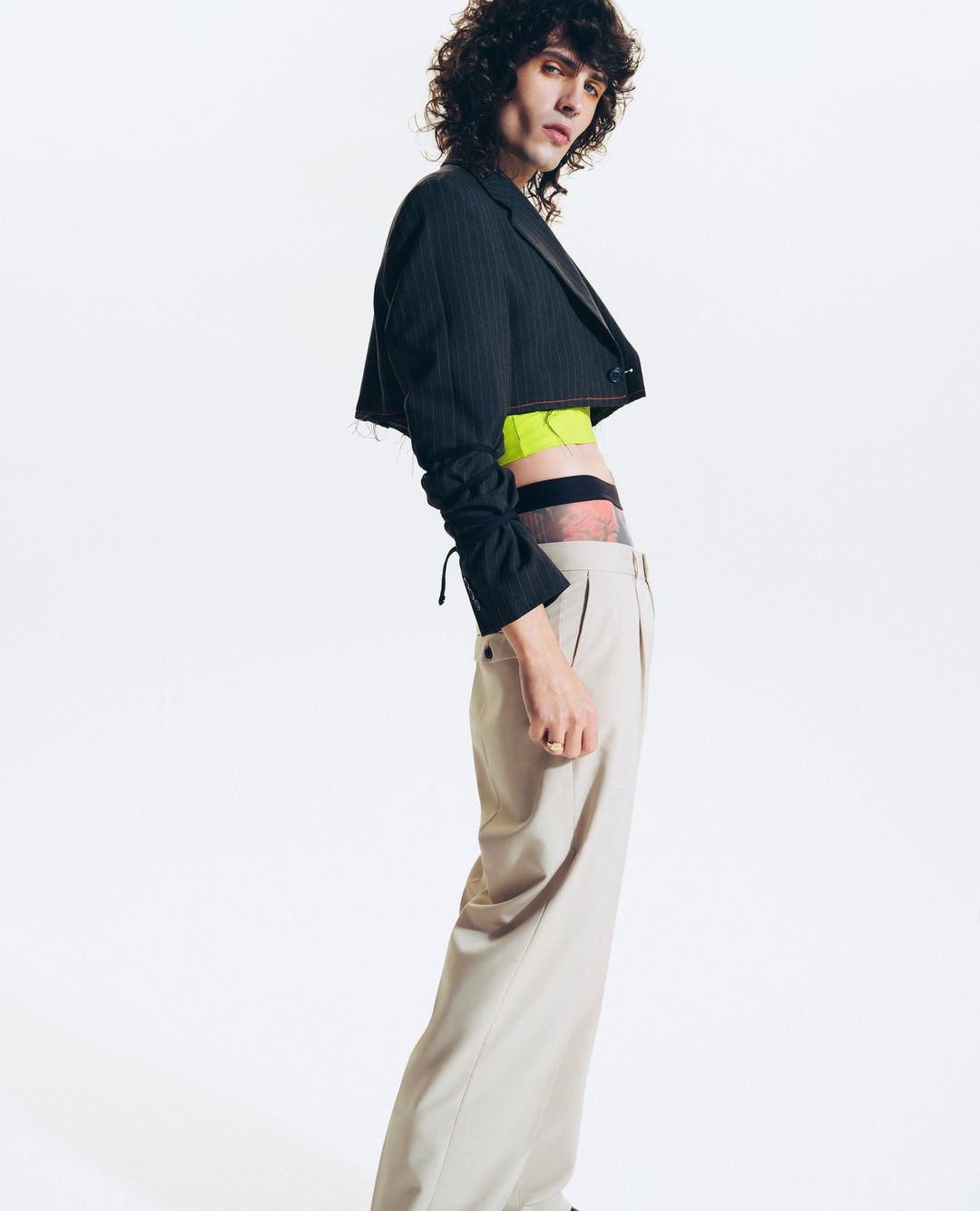 An adult male with curly dark hair poses in a photo studio wearing a black cropped jacket and beige trousers against a white backdrop.