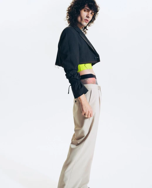 An adult male with curly dark hair poses in a photo studio wearing a black cropped jacket and beige trousers against a white backdrop.