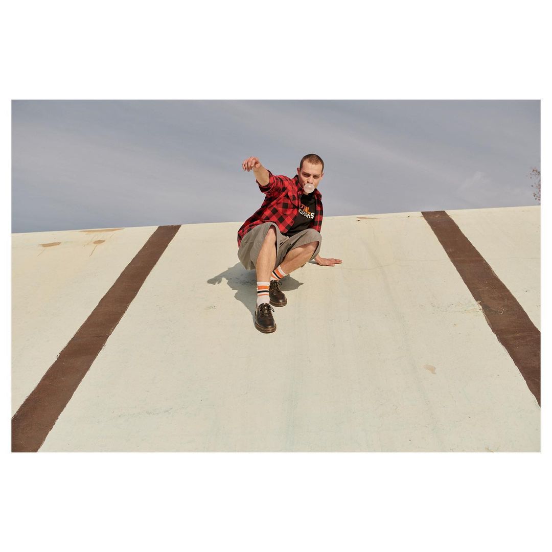 Man crouches on a white surface with brown lines, blowing a bubble with gum. He wears a red and black flannel shirt and shorts.