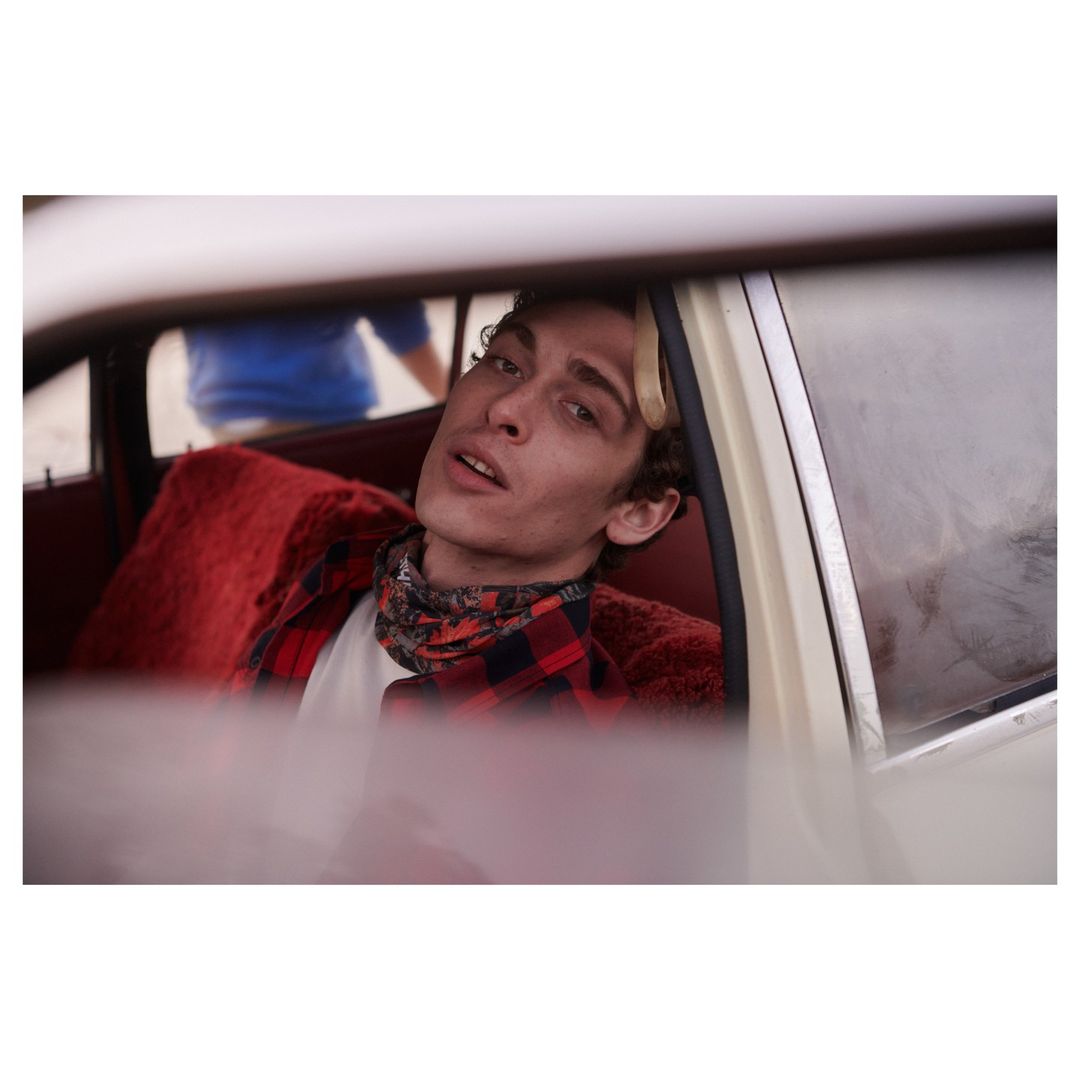 Young adult man in a red flannel shirt sits in a vintage car with red upholstery, looking at the camera, in a medium shot.