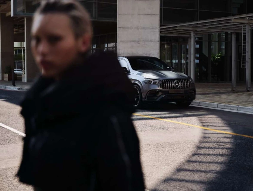 Gray Mercedes-Benz GLE parked on a city street with an adult in a black coat passing by, modern building in the background.