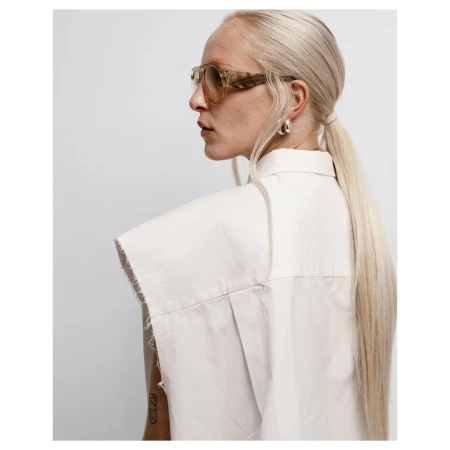Profile portrait of a blonde woman wearing tortoiseshell sunglasses and a white sleeveless shirt against a plain background