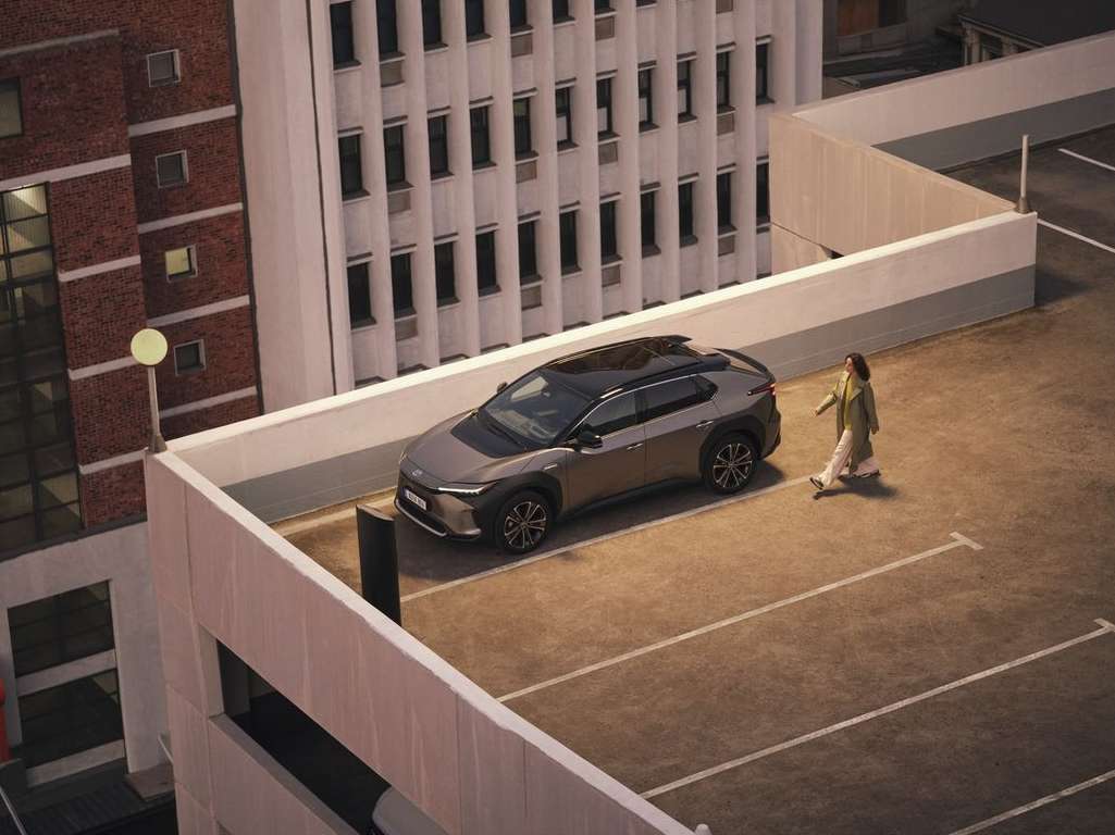 A gray SUV is parked on a rooftop parking lot beside a woman walking. Tall buildings are in the background on a cloudy day.