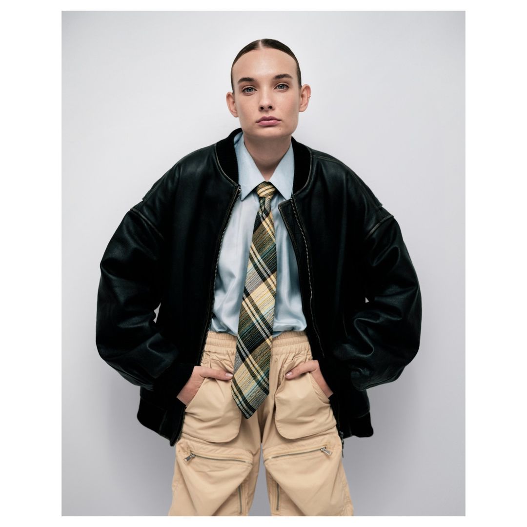 Model in black leather bomber jacket, light blue shirt, plaid tie, and beige cargo pants against a white background.