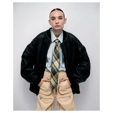 Model in black leather bomber jacket, light blue shirt, plaid tie, and beige cargo pants against a white background.