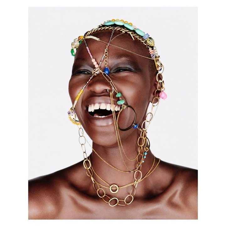 Close-up of a smiling African woman adorned with colorful beaded jewelry around her head and neck against a plain white background.