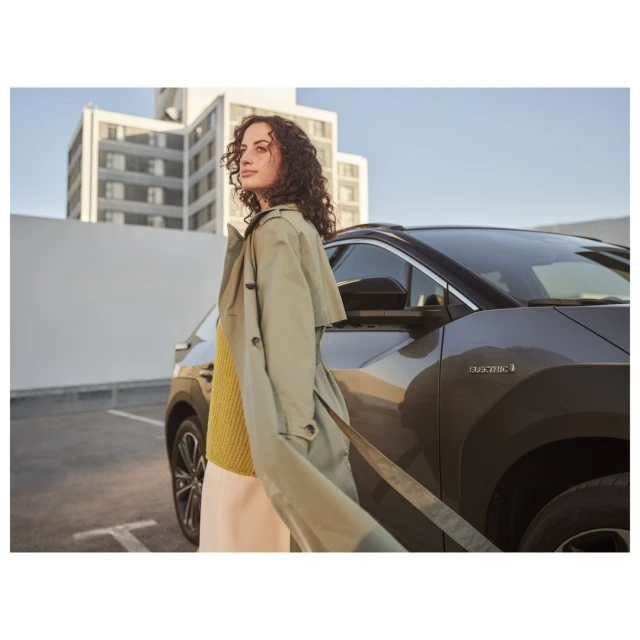 A woman in a tan coat stands near a dark grey car with a modern building in the background on a sunny day in an urban setting.