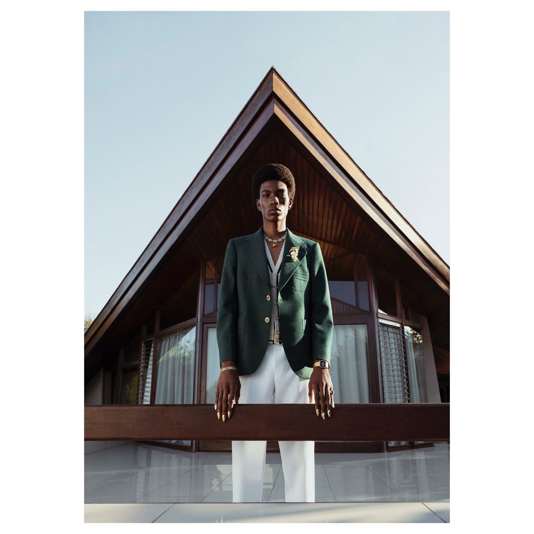 Fashionable adult male poses in a green blazer and white pants, standing outdoors in front of modern triangular architecture.