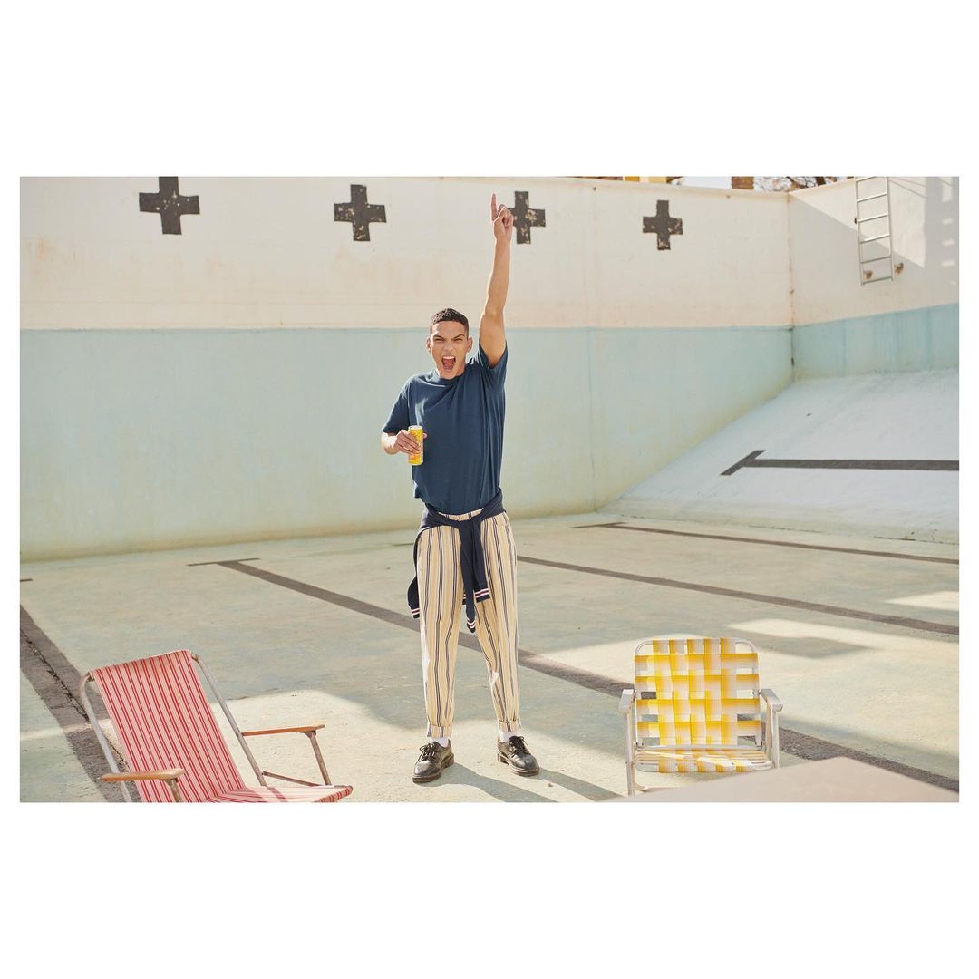 Man standing in empty pool holding a drink, wearing a t-shirt and striped pants, with beach chairs visible in the shot.