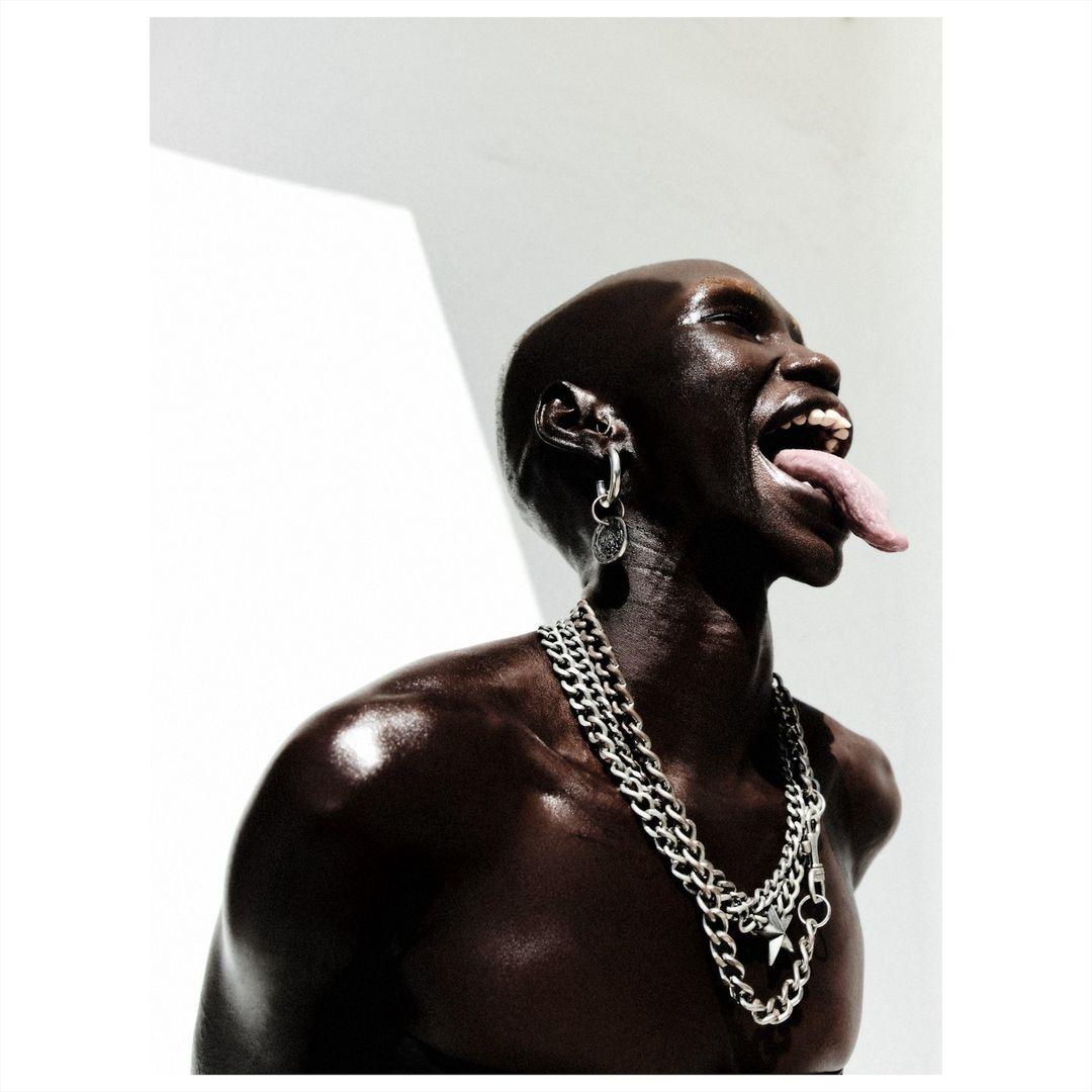 Close-up studio shot of a shiny, dark-skinned bald adult with tongue out, wearing silver jewelry and a chain necklace against a white background.