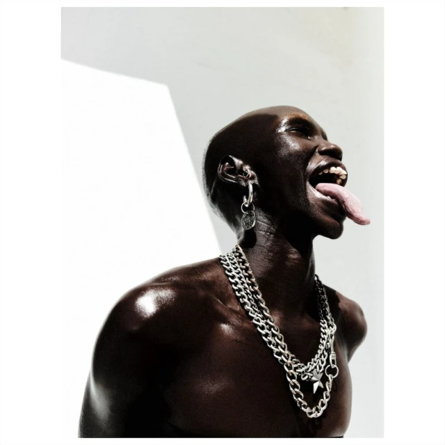 Close-up studio shot of a shiny, dark-skinned bald adult with tongue out, wearing silver jewelry and a chain necklace against a white background.