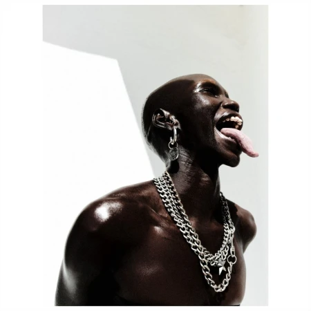 Close-up studio shot of a shiny, dark-skinned bald adult with tongue out, wearing silver jewelry and a chain necklace against a white background.