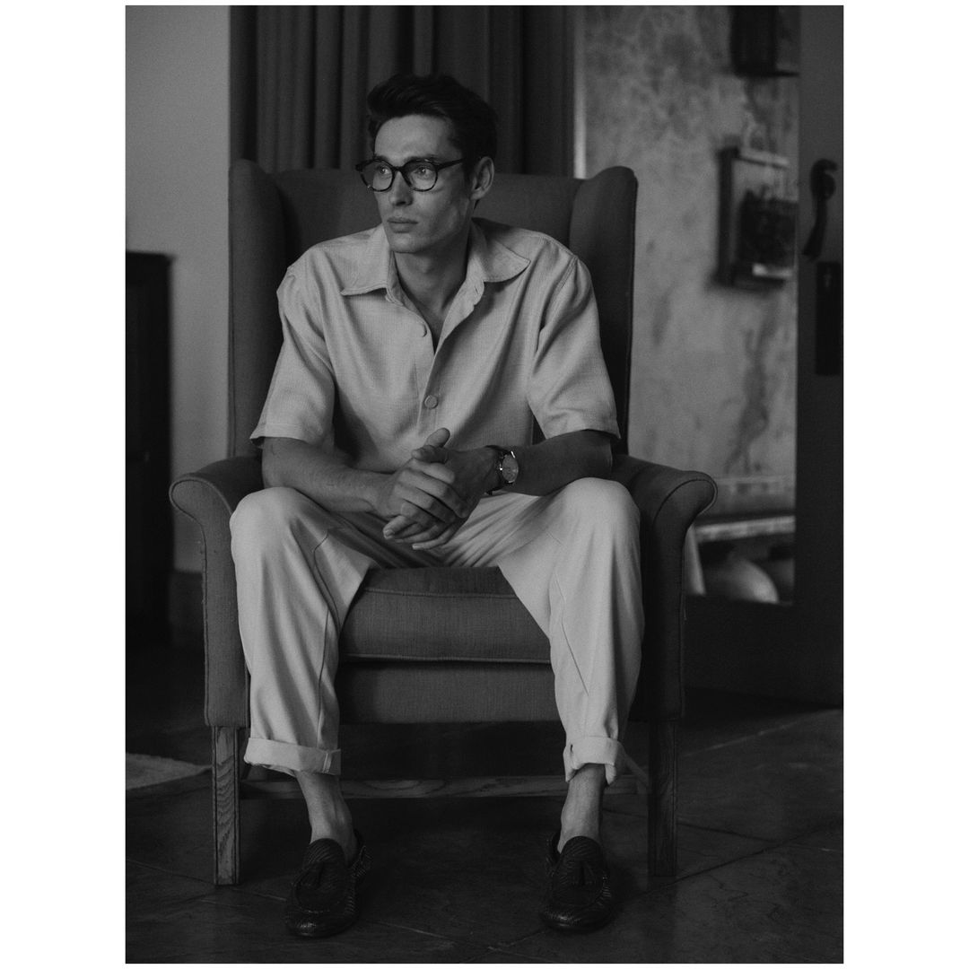 Black and white shot of a man with glasses sitting in a wingback chair, wearing a short-sleeved shirt, pants, and loafers.