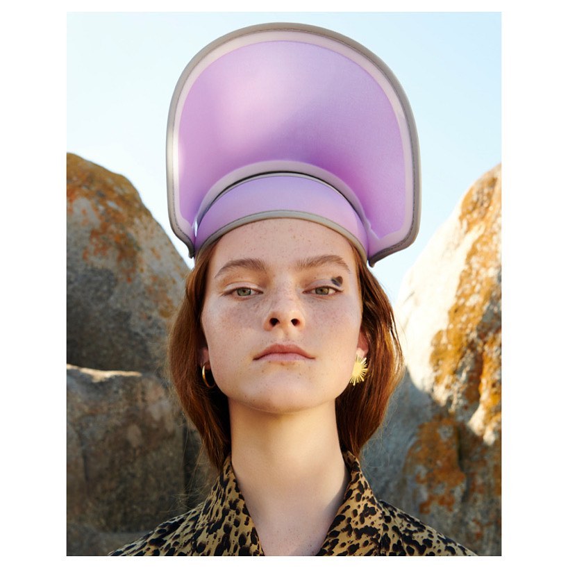 Close-up of a young woman wearing a sculptural lilac hat and leopard print shirt, posing in an outdoor setting with rocks.
