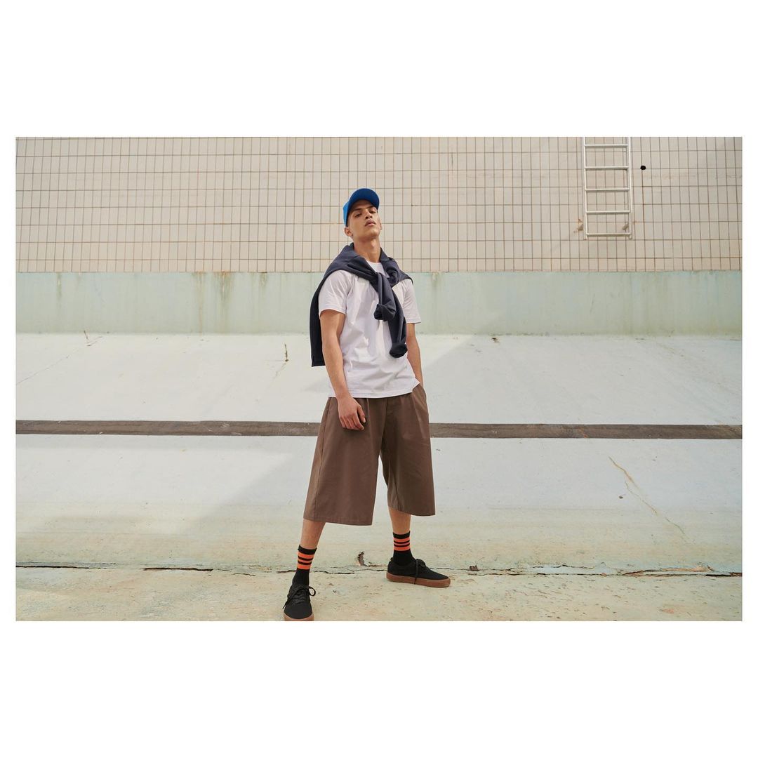 Man in white t-shirt, blue cap, and brown shorts stands against a textured wall, a navy sweater draped over his shoulders.