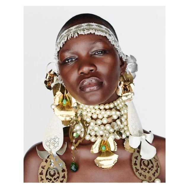 Close-up portrait of a striking dark-skinned woman with intricate beaded headband and pearl necklace in a bright photo studio setting.