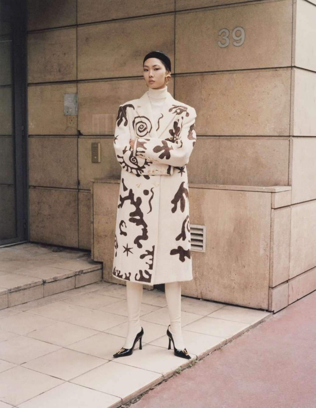 Fashion shot of a stylish adult woman in a patterned coat and cream-colored leggings posing in front of a tan building with the number 39.