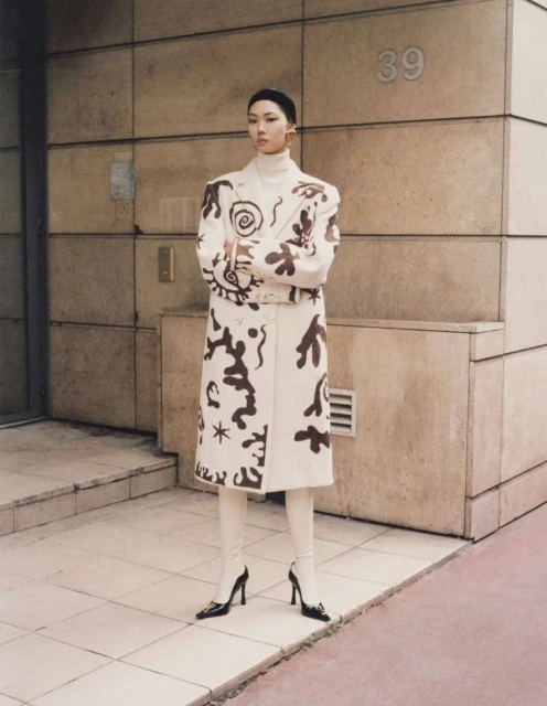 Fashion shot of a stylish adult woman in a patterned coat and cream-colored leggings posing in front of a tan building with the number 39.
