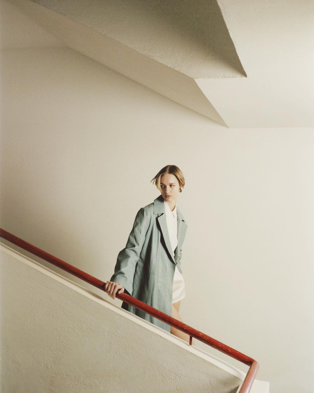 Fashion shot of an adult female with blonde hair wearing a green coat and a white top, posing on stairs with a red handrail.
