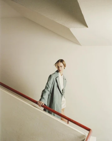 Fashion shot of an adult female with blonde hair wearing a green coat and a white top, posing on stairs with a red handrail.