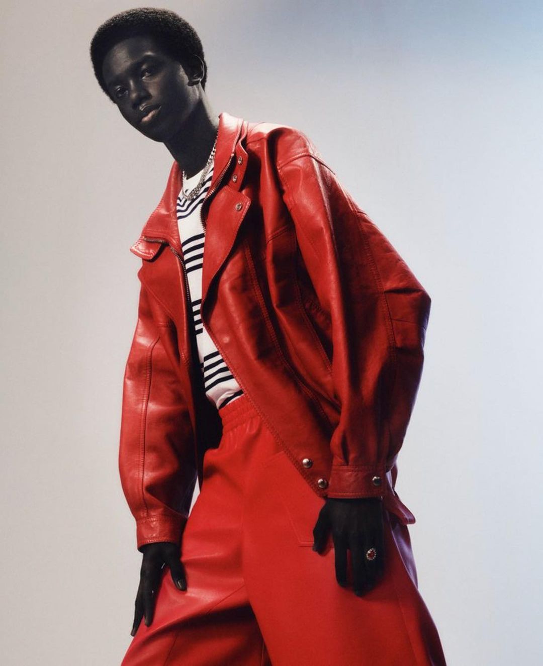 Fashion shot of a single Black male model wearing a red leather jacket and matching pants, posing against a gradient background.