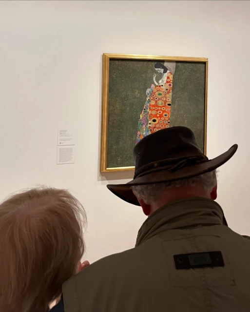Two adults view a painting in an art gallery; the artwork features a figure in an orange patterned robe, displayed in a gold frame.