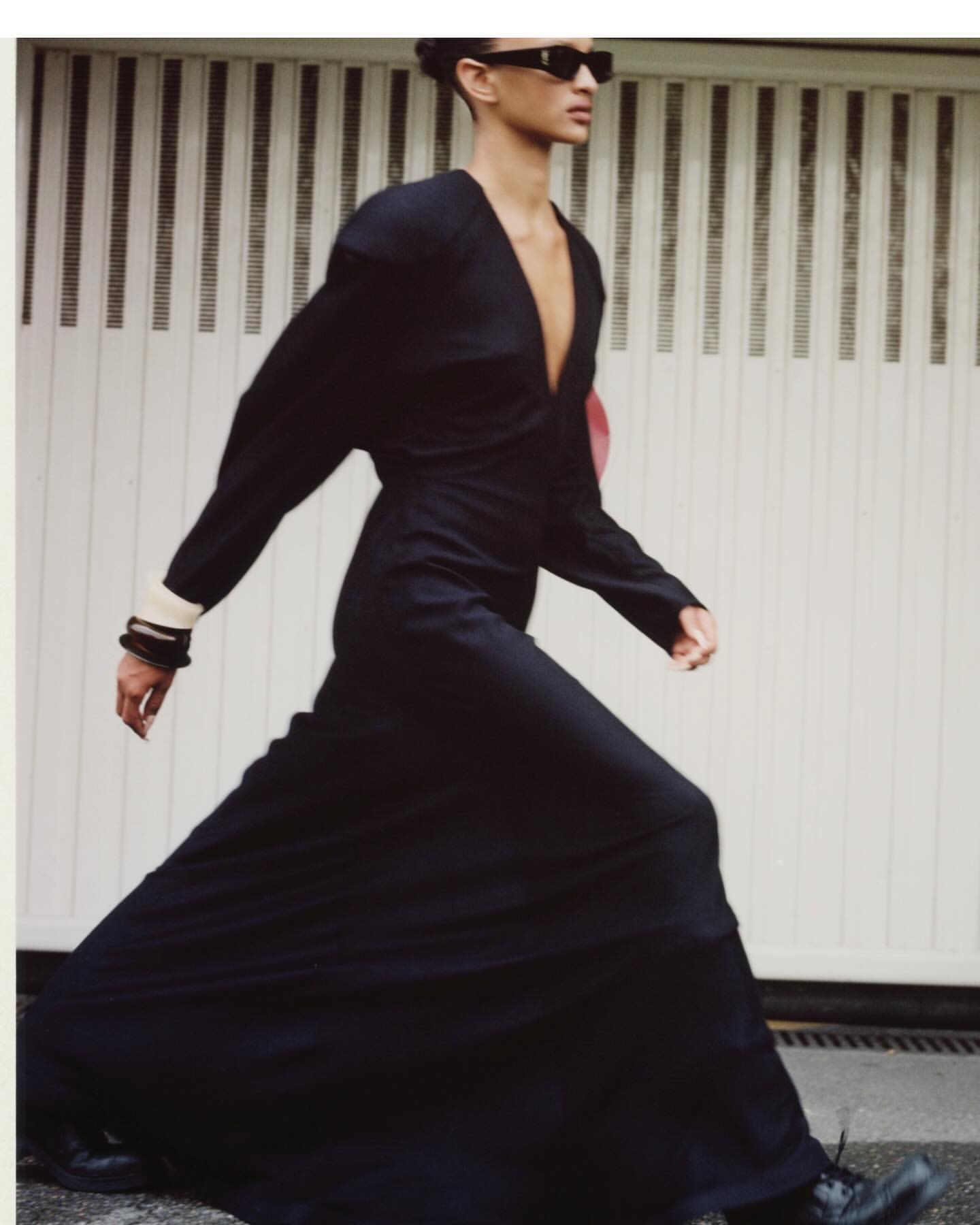 Model in a long black dress and sunglasses walking in front of a white garage door by Antonio Dicorato