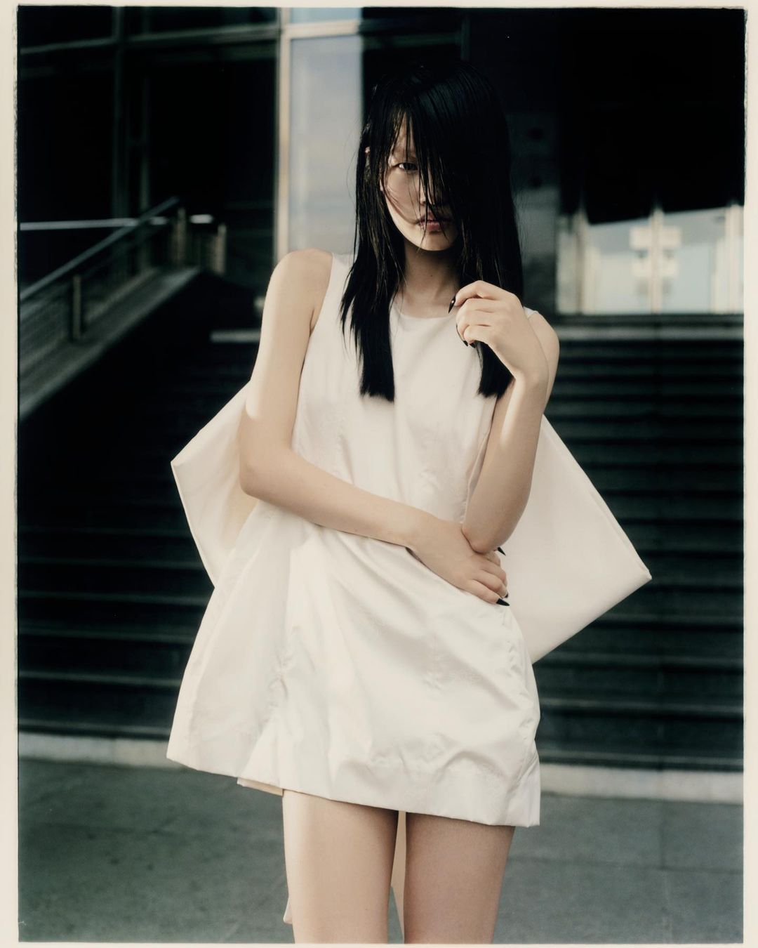 Young woman in a sleeveless mini dress posing outdoors against a modern staircase and glass-front building, editorial fashion shot.