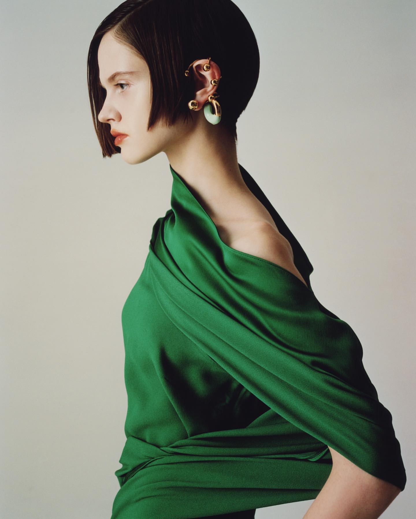 A young, elegant woman with a bob haircut poses in a profile shot, wearing a green gown and ear cuff in a brightly lit, neutral studio.