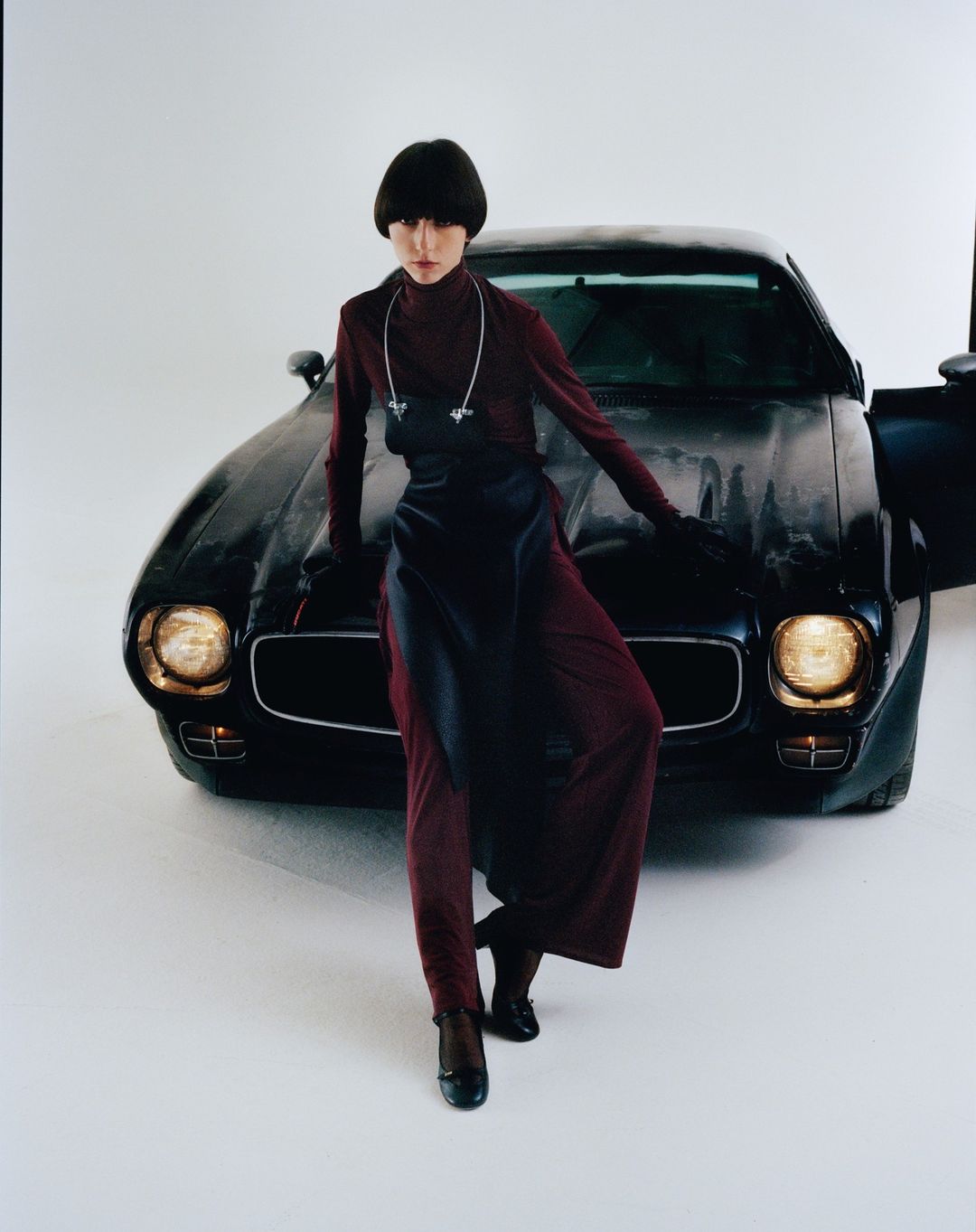 Fashionable woman in maroon outfit poses with classic black car in a studio setting with a light background, striking a confident pose.