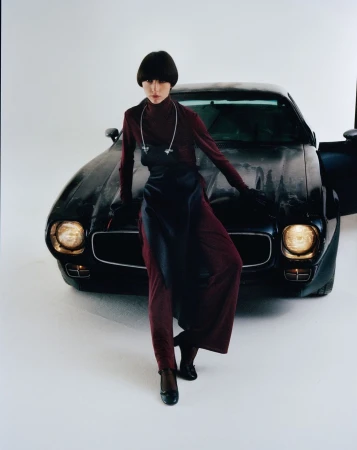 Fashionable woman in maroon outfit poses with classic black car in a studio setting with a light background, striking a confident pose.