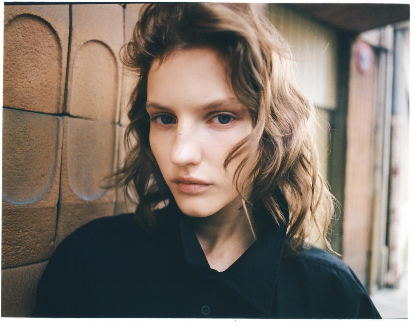 Close-up portrait of a young adult woman with wavy brown hair, blue eyes, wearing dark clothing against a textured brown wall outdoors.
