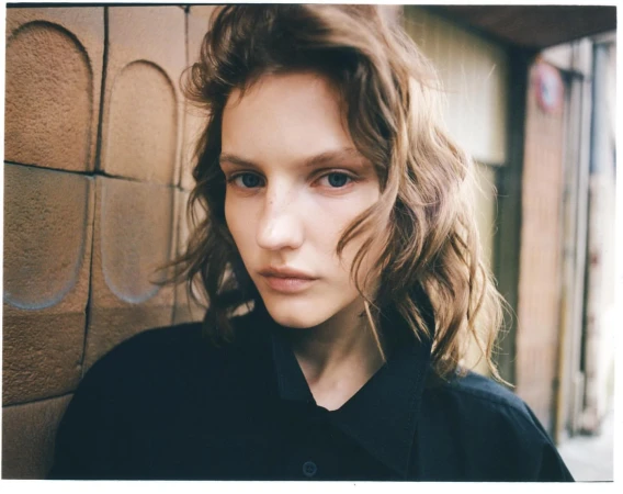 Close-up portrait of a young adult woman with wavy brown hair, blue eyes, wearing dark clothing against a textured brown wall outdoors.
