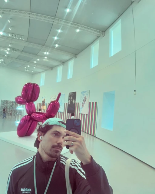Man takes a selfie at an art gallery with a pink balloon animal sculpture in the background, wearing a baseball cap and athletic jacket.