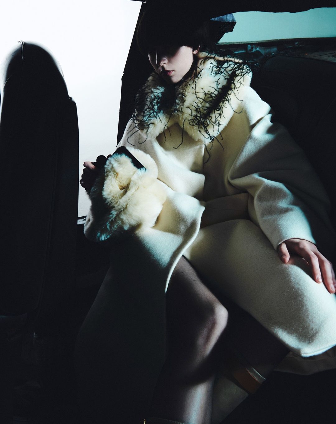 Woman in a cream-colored coat with fur trim poses indoors with dark lighting. Fashion editorial portrait.