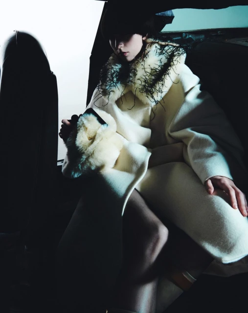 Woman in a cream-colored coat with fur trim poses indoors with dark lighting. Fashion editorial portrait.