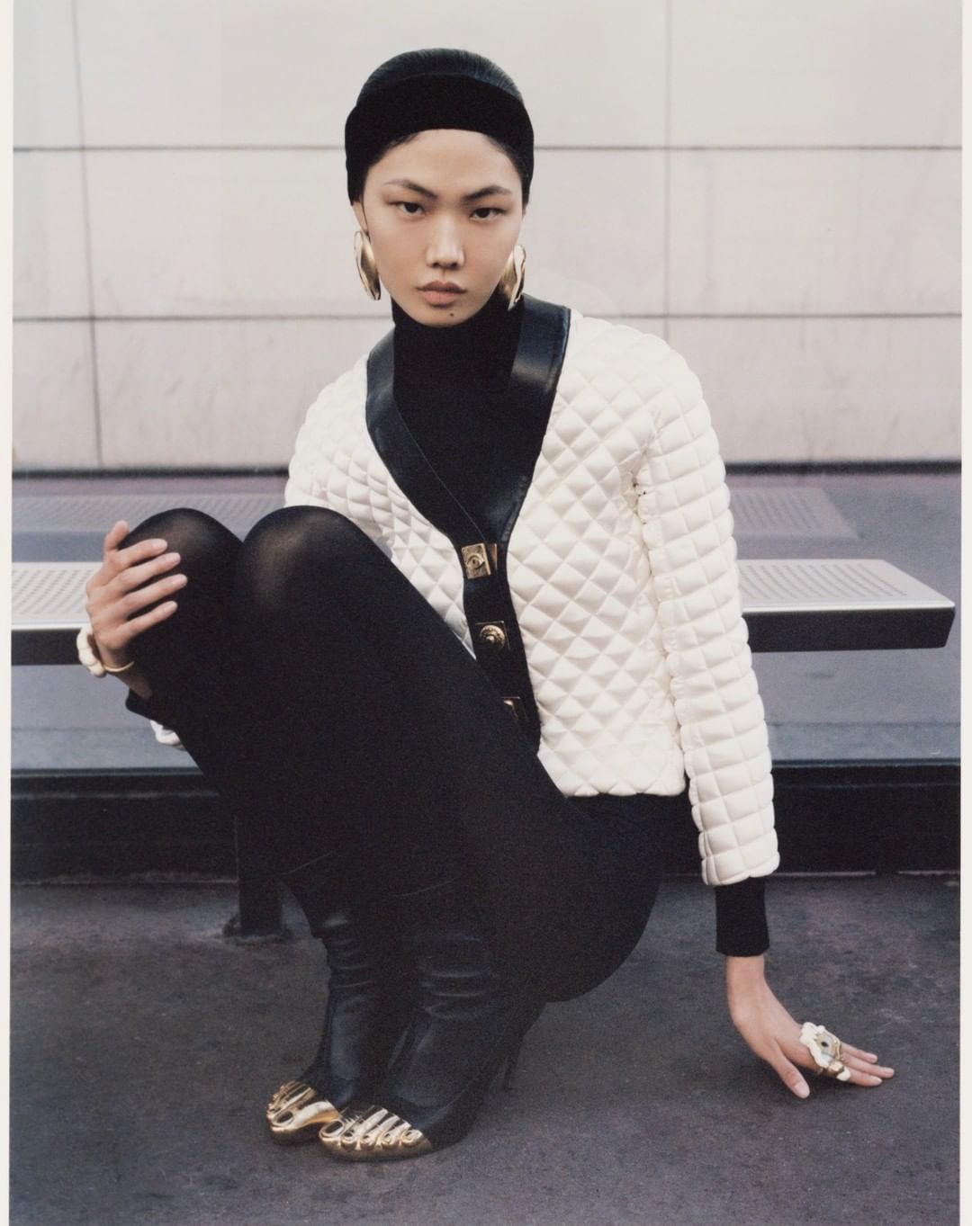 Asian model in a black turtleneck and leggings, wearing a white quilted cardigan with gold-tipped shoes, sits in a fashion editorial.