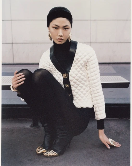 Asian model in a black turtleneck and leggings, wearing a white quilted cardigan with gold-tipped shoes, sits in a fashion editorial.