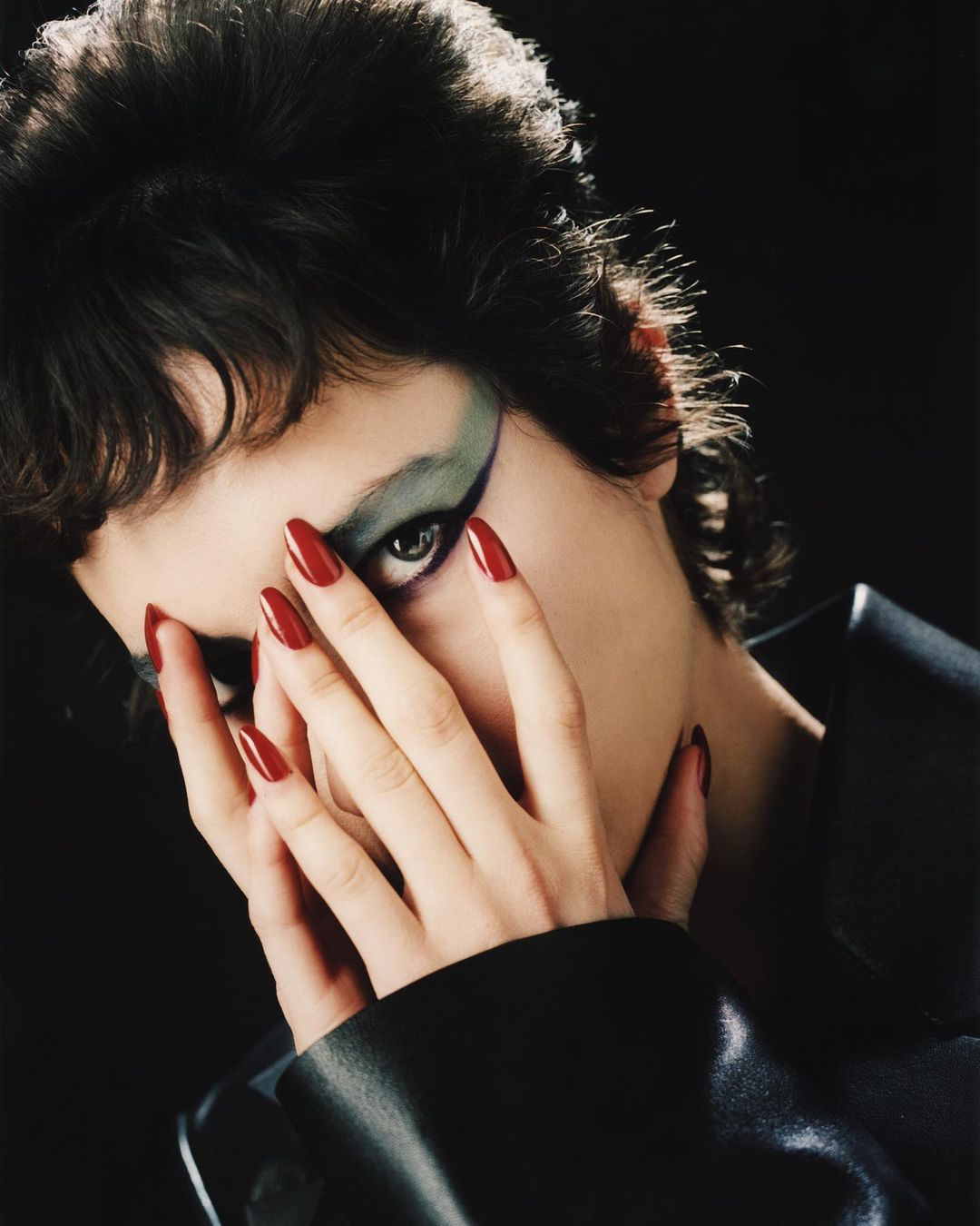 Portrait of a woman with dramatic makeup and long red nails posing in front of a dark background in a beauty fashion shot.