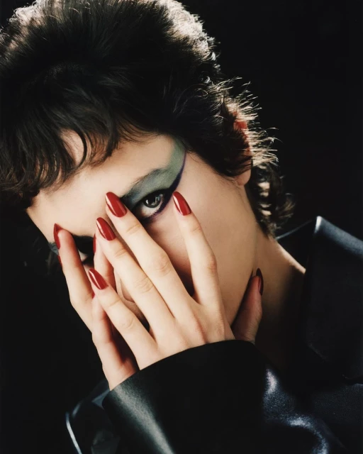 Portrait of a woman with dramatic makeup and long red nails posing in front of a dark background in a beauty fashion shot.