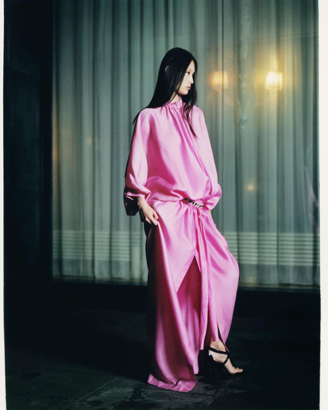 Elegant Asian woman in a pink satin gown and black strappy high heels. Soft window lighting and modern fashion style.