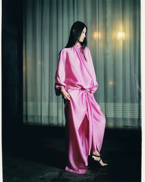 Elegant Asian woman in a pink satin gown and black strappy high heels. Soft window lighting and modern fashion style.